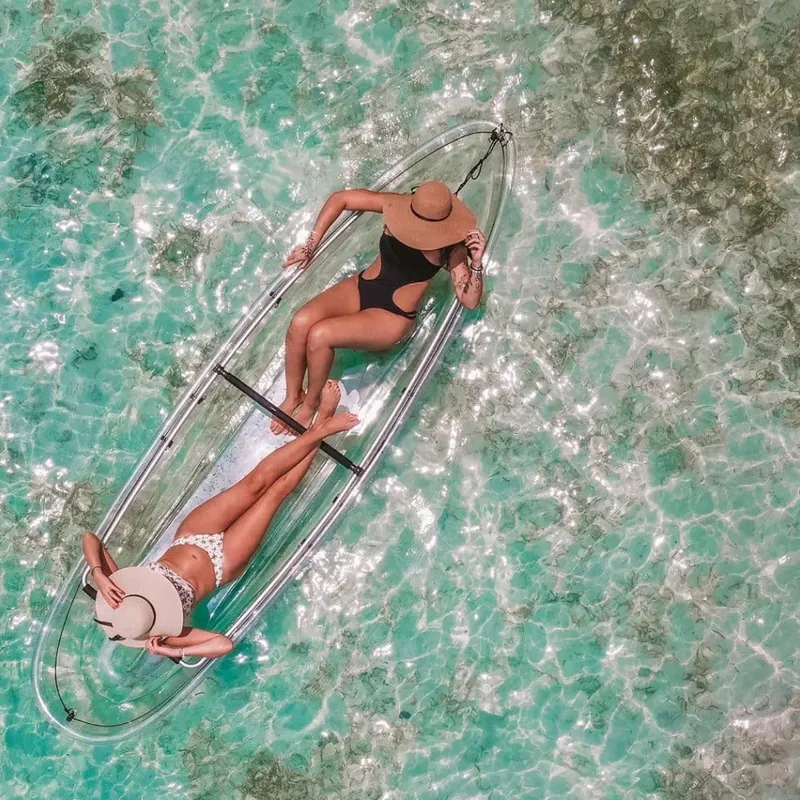 Guest in a sun hat lying in a clear kayak above rippled turquoise shallows
